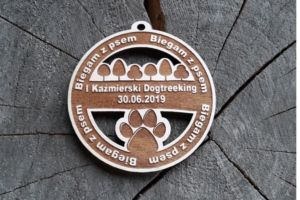 Okrągły ażurowy medal drewniany z wyciętym laserowo motywem łapy psa, napisem I Kaźmierski Dogtreeeking oraz wyciętym ażurowo zarysem drzew.