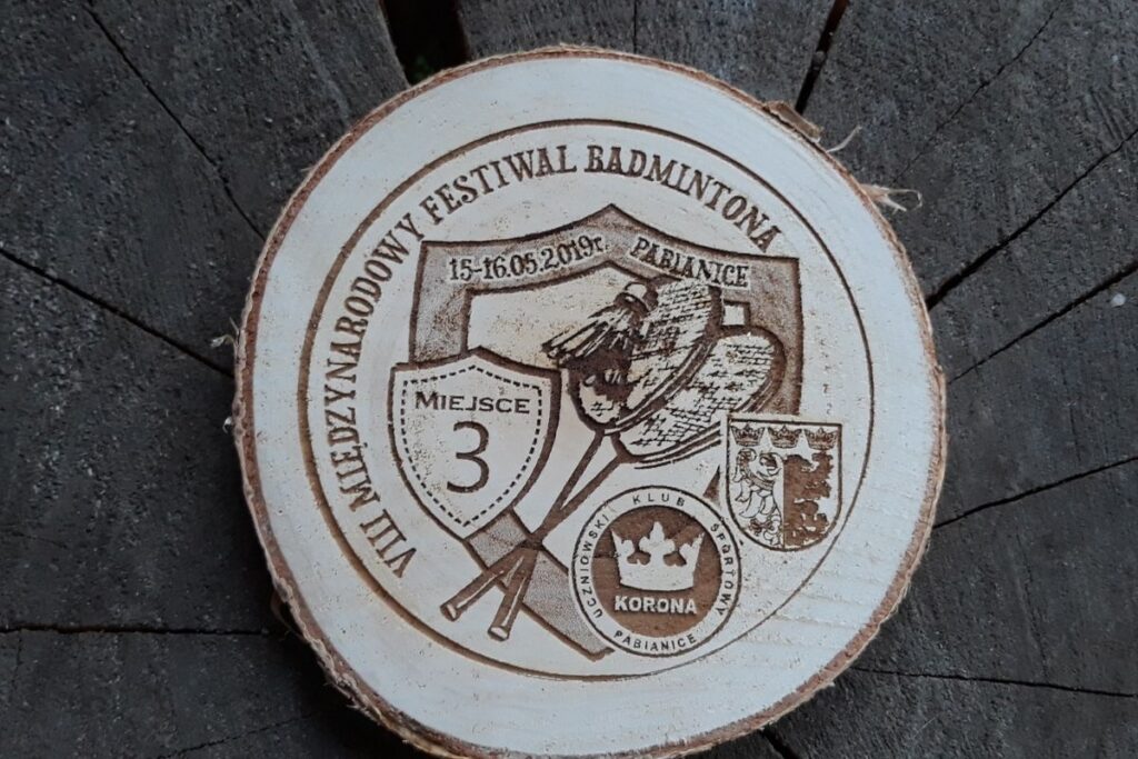 Medal wykonany z naturalnego plastra brzozy z korą, z grawerem lotki do badmintona, rakiety i napisem Festiwal Badmintona.