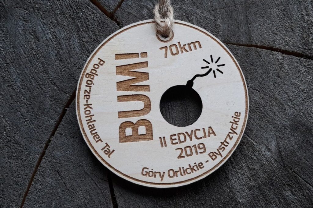 Okrągły, drewniany medal w kształcie koła z wycięciem na wylot komiksowej bomby z palącym się lontem i napisem BUM.