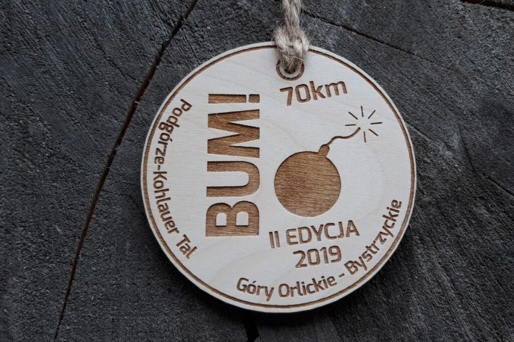 Okrągły, drewniany medal w kształcie koła z grawerem komiksowej bomby z palącym się lontem i napisem BUM.