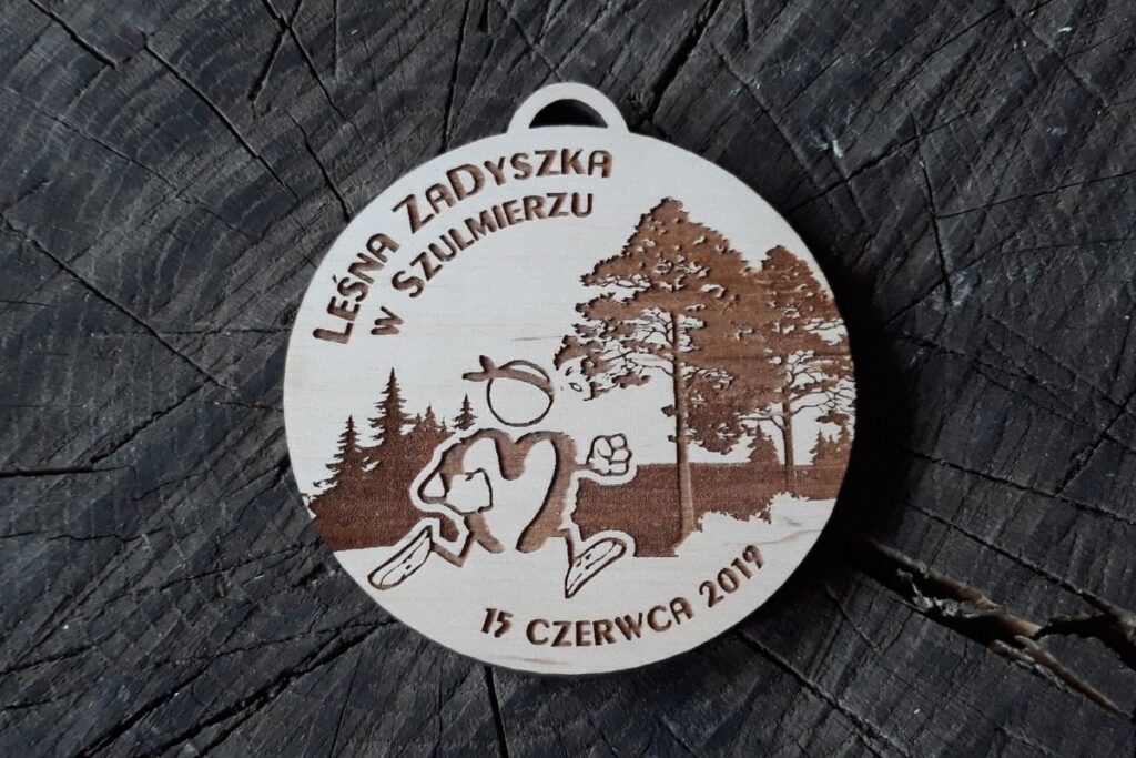 Okrągły drewniany medal z grawerem gęstego lasu iglastego, sylwetką biegacza w formie loga w ruchu oraz napisem Leśna Zadyszka.