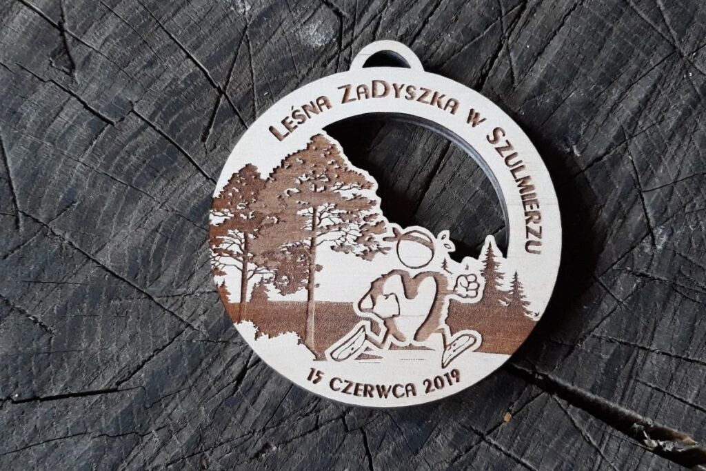 Okrągły drewniany medal z grawerem gęstego lasu iglastego, sylwetką biegacza w formie loga w ruchu oraz napisem Leśna Zadyszka.