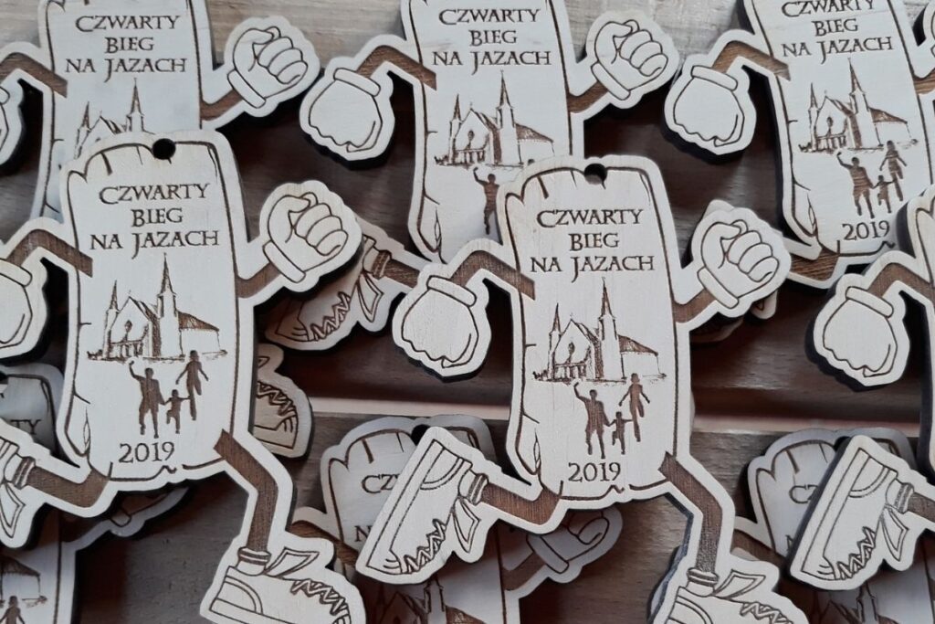 Drewniany medal w kształcie kłody drewna z wyrastającymi nogami w ruchu biegowym, napisem IV Bieg na Jazach i widocznymi słojami.