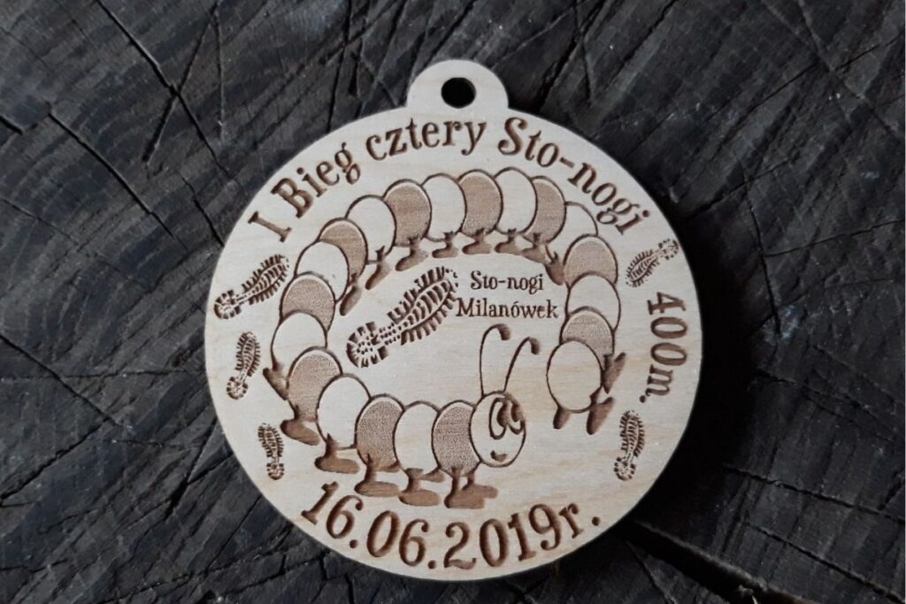 Okrągły drewniany medal z grawerem uśmiechniętej stonogi w butach biegowych, napisem I Bieg Cztery-Sto-Nogi i datą wydarzenia.