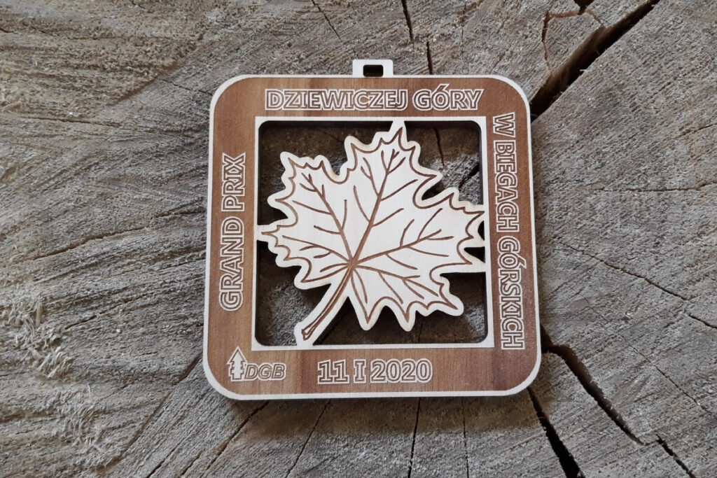 Kwadratowy medal drewniany z ażurowym wycięciem w kształcie liścia klonu i grawerowanym napisem na dolnej krawędzi.