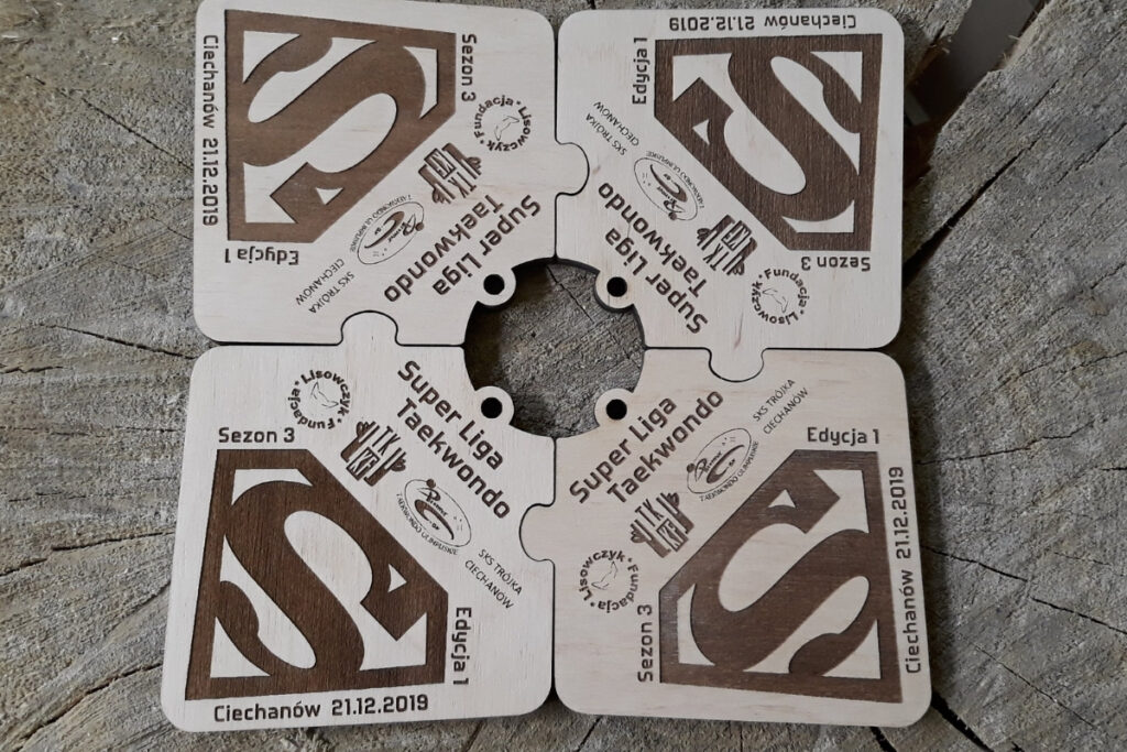 Cztery drewniane medale o nieregularnych kształtach, które po złożeniu pasują do siebie jak puzzle, tworząc zbliżoną do kwadratu tabliczkę z pełną grafiką.