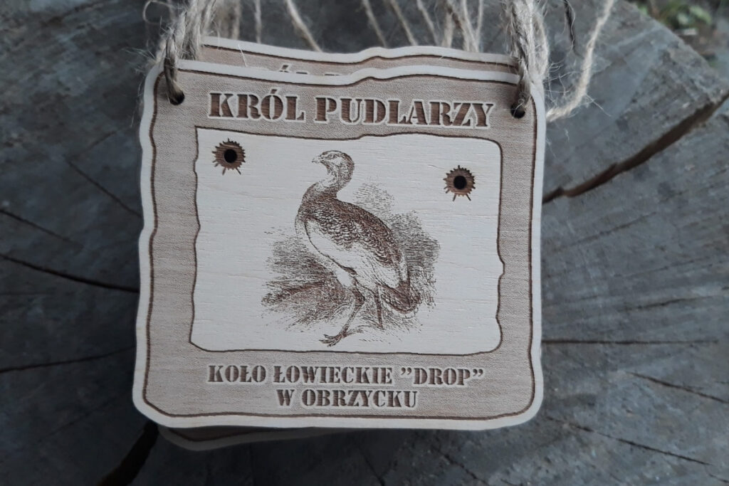 Prostokątny drewniany medal z grawerem dropia, napisem Król Pudlarzy oraz kulami - wycięciami na wylot.