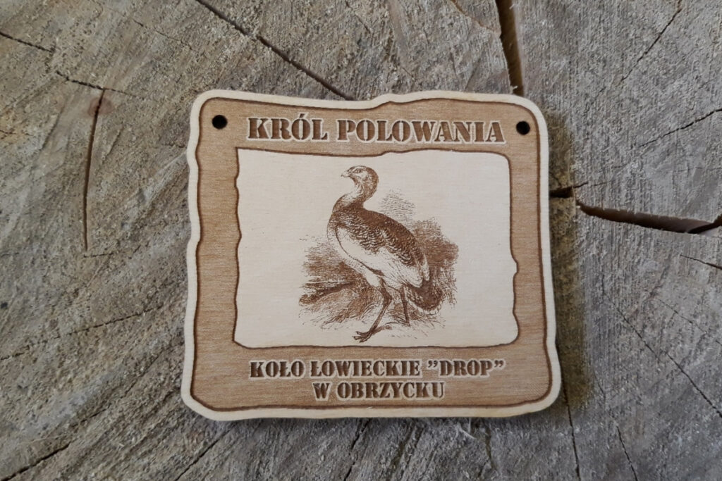 Prostokątny drewniany medal z grawerem dropia w dumnej postawie, napisem Król Polowania oraz datą wydarzenia.