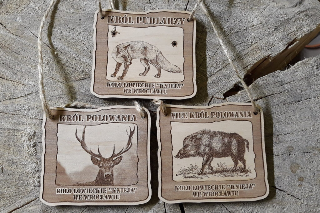 Zestaw trzech drewnianych medali łowieckich: Król Polowania z jeleniem, Vicekról z dzikiem oraz Król Pudlarzy z lisem, wykonane z grawerowanej sklejki z brzozy.