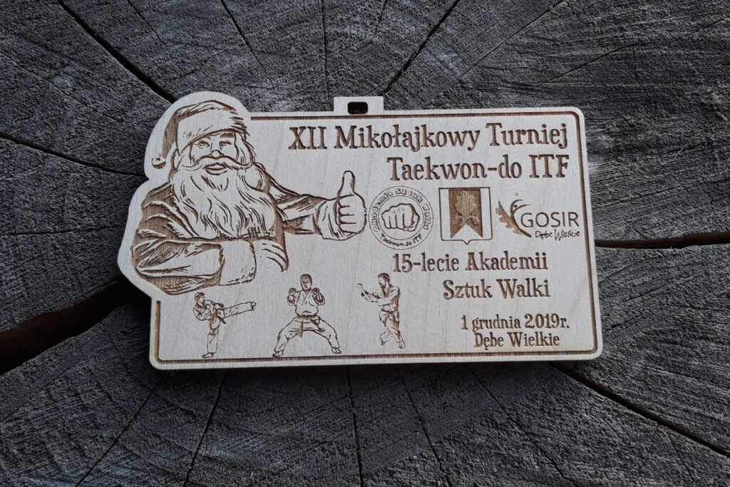 Drewniany medal z grawerem radosnej twarzy Mikołaja w czapce, napisem Mikołajkowy Turniej Taekwon-do oraz logotypami.