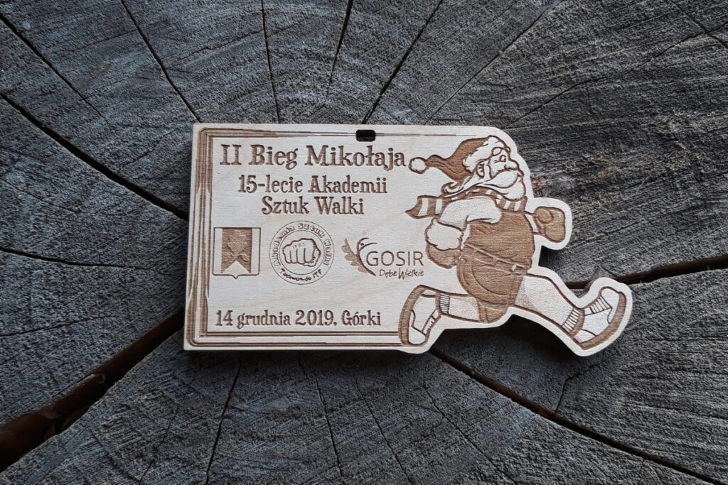 Drewniany medal z grawerem radosnej twarzy Mikołaja w czapce, napisem Bieg Mikołaja oraz logotypami.