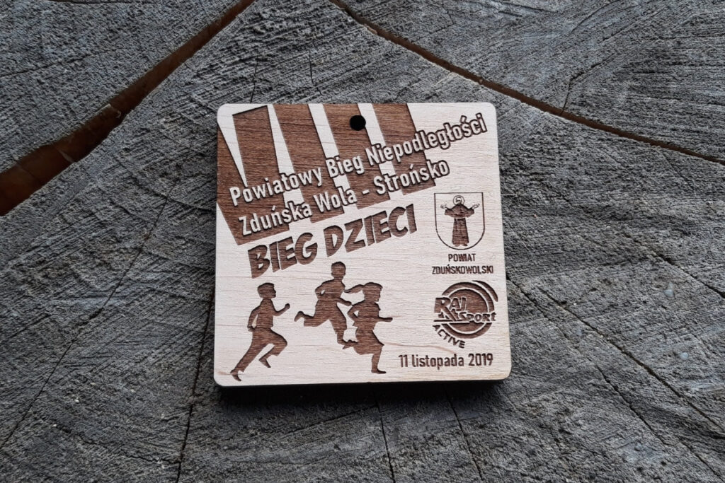 Drewniany, kwadratowy medal z napisami: Powiatowy Bieg Niepodległości - VII Bieg Dzieci, logotypami i biegnącymi dziećmi.