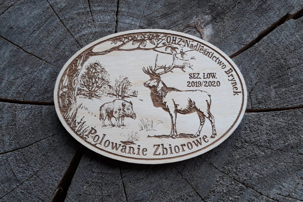 Owalny drewniany medal myśliwski z grawerem jelenia byka oraz dzika, ozdobiony drzewami.