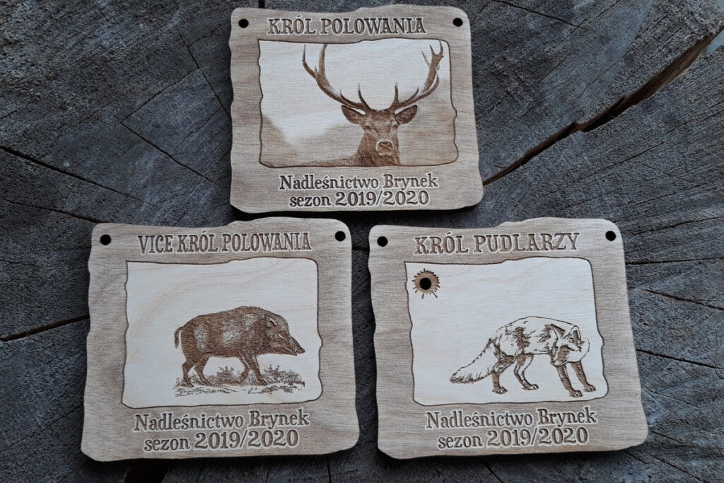 Zestaw trzech prostokątnych medali drewnianych z grawerami: jeleń (Król), dzik (Vicekról) i lis (Król Pudlarzy) na jasnej sklejce brzozowej.