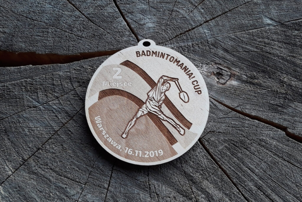 Okrągły drewniany medal do badmintona z grawerem gracza, napisem 'Badmintomania! Cup' i dekoracyjnymi łukami symbolizującymi ruch.