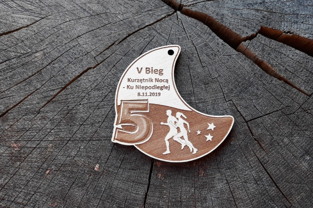 Drewniany medal w kształcie półksiężyca z grawerem nocnego nieba, gwiazd, sylwetek biegaczy i napisami.