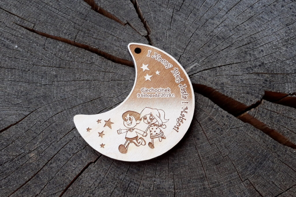 Drewniany medal w formie półksiężyca z grawerem dziecięcych sylwetek Jasia i Małgosi na tle lasu i gwiazd, z napisem Nocny Bieg.