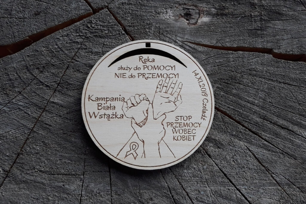 Okrągły drewniany medal z grawerem splecionych dłoni, napisem Stop Przemocy Wobec Kobiet oraz symbolem białej wstążki.