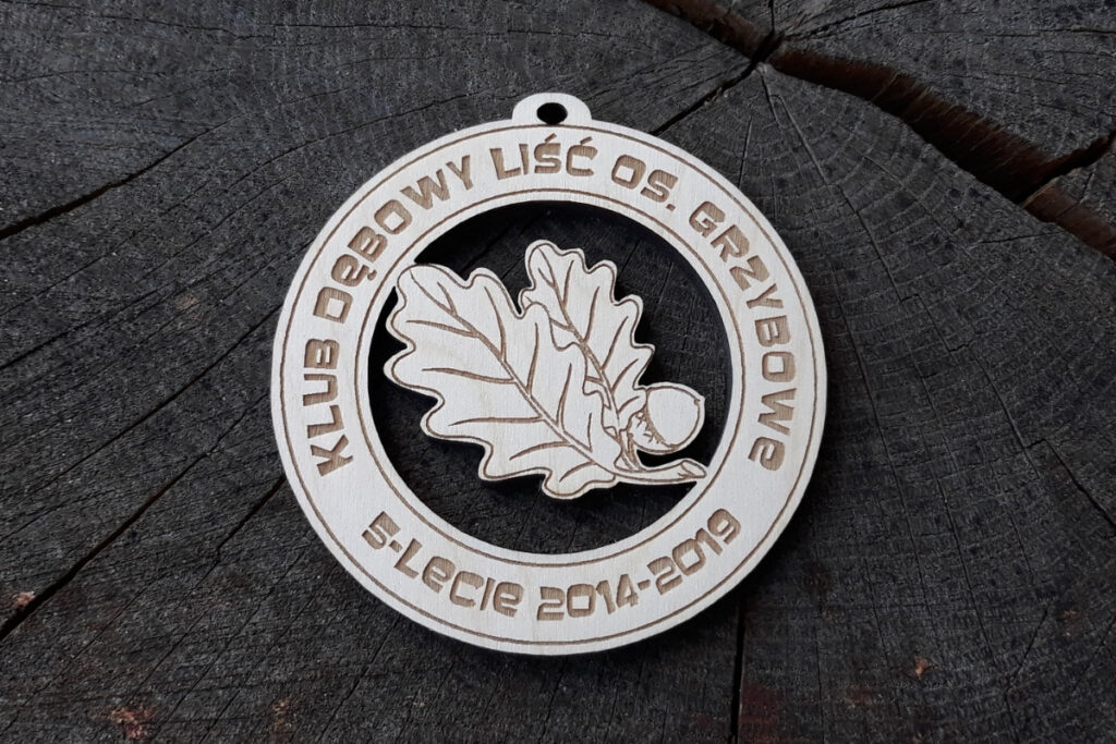 Okrągły drewniany medal z ażurowym wycięciem w kształcie liścia dębu, wykonany z jasnej brzozy, z grawerowanymi napisami po okręgu.