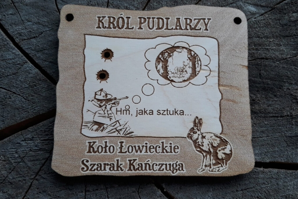 Prostokątny drewniany medal dla Króla Pudlarzy z grawerem myśliwego celującego do jelenia, który ostatecznie trafia obok jelenia.
