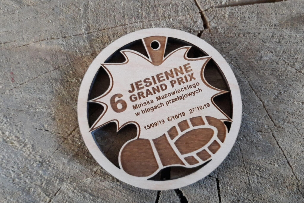 Okrągły drewniany medal z ażurowym wycięciem w kształcie liścia dębu w centralnej części i grawerowanym napisem wokół krawędzi.