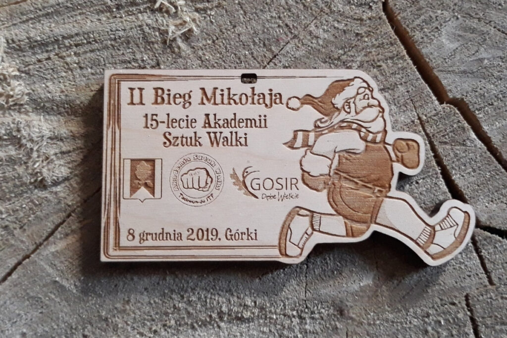 Drewniany medal z grawerem uśmiechniętej buzi Mikołaja w czapce z pomponem i napisem II Bieg Mikołaja