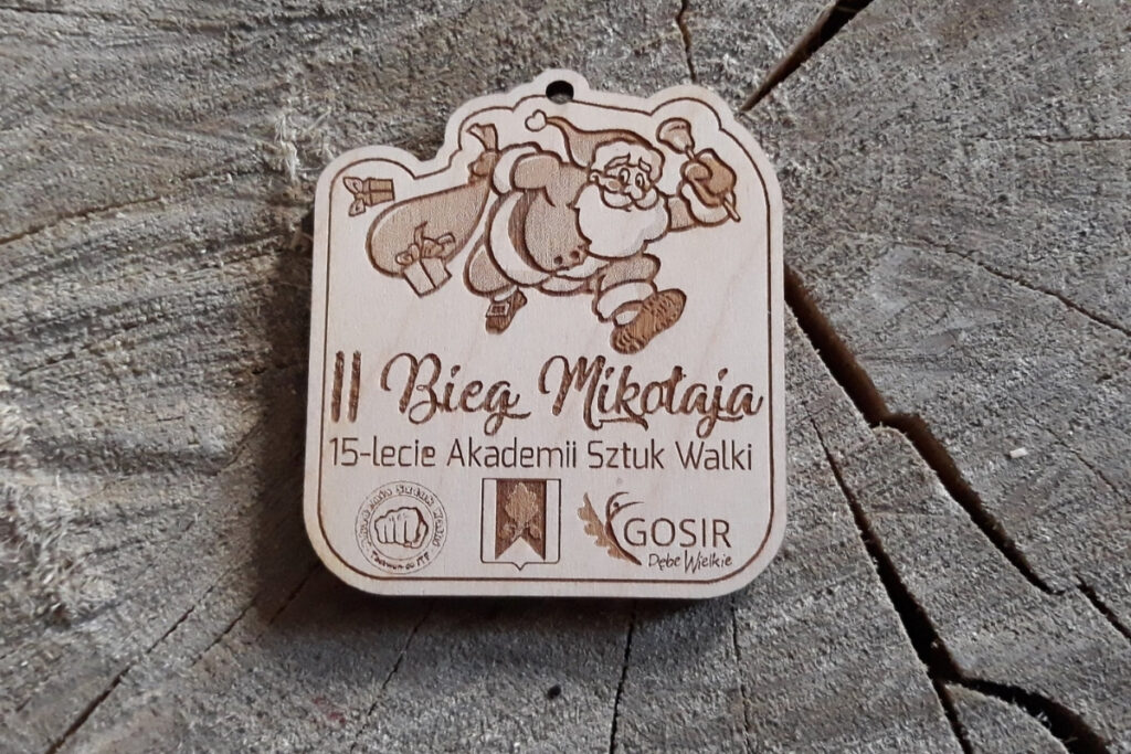 Drewniany medal z grawerem uśmiechniętej buzi Mikołaja w czapce z pomponem i napisem II Bieg Mikołaja