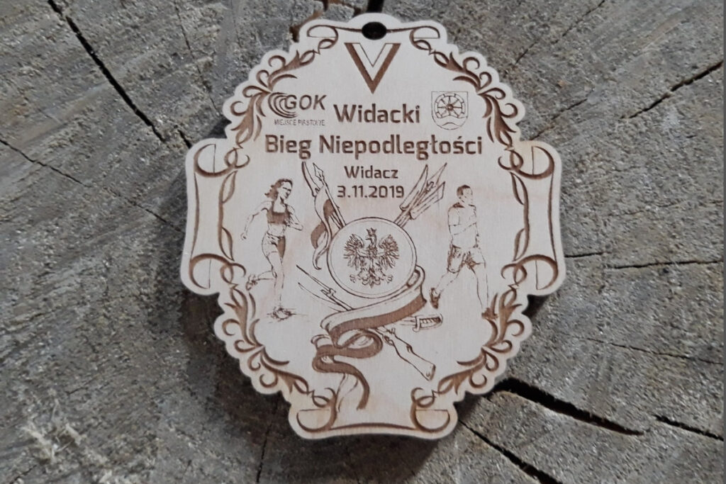 Drewniany medal nieregularnych kształtów z napisem V Widacki Bieg Niepodległości i elementami patriotycznymi - biało czerwoną wstążką, orłem itp.