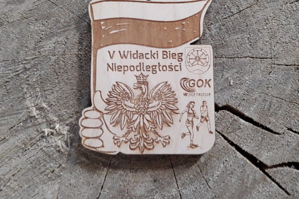 Drewniany medal nieregularnych kształtów z napisem V Widacki Bieg Niepodległości i elementami patriotycznymi - biało czerwoną wstążką, orłem itp.