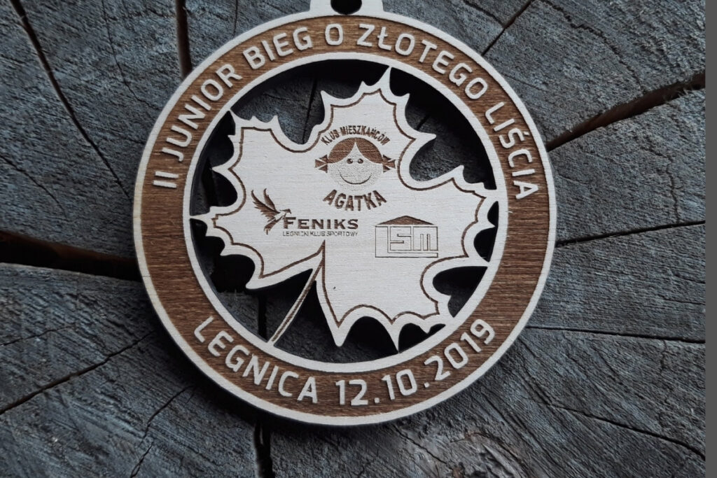 Okrągły drewniany medal dla dzieci z ażurowo wyciętym liściem w centralnej części i grawerowanym napisem Bieg Juniorów o Złotego Liścia.