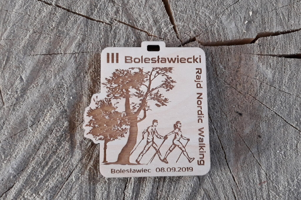 Drewniany medal z grawerem sylwetki maszerującej osoby z kijami Nordic Walking, napisem III Bolesławiecki Rajd i elementami roślinnymi.
