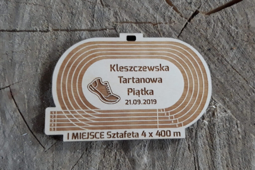 Drewniany medal w kształcie owalnej bieżni lekkoatletycznej z wygrawerowanymi torami, napisem oraz grafiką buta.