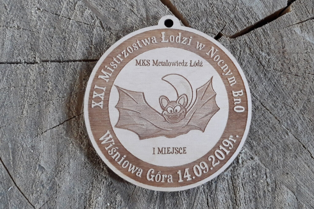 Okrągły drewniany medal z grawerem nietoperza z rozpostartymi skrzydłami, napisem XXI Mistrzostwa Łodzi w Nocnym BnO i logo organizatora.
