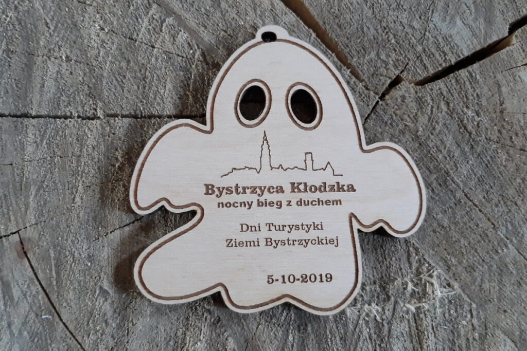 Drewniany medal wycięty w kształcie ducha z grawerem Nocny Bieg z Duchem, rycin miasta i wyciętymi ażurowo na wylot oczami.
