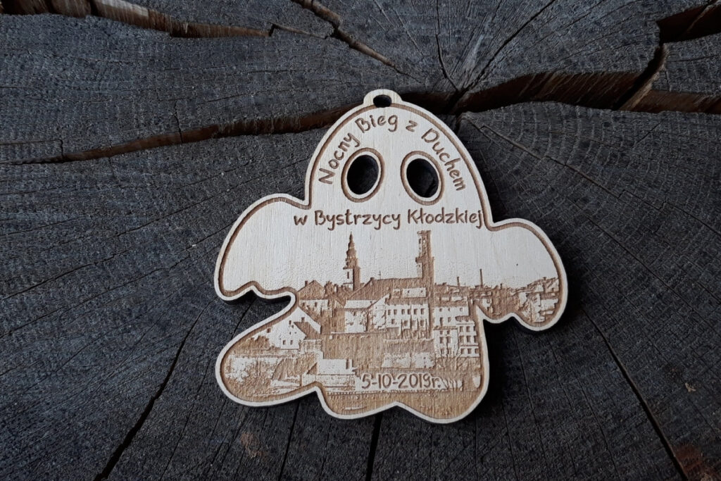 Drewniany medal wycięty w kształcie ducha z grawerem Nocny Bieg z Duchem, rycin miasta i wyciętymi ażurowo na wylot oczami.