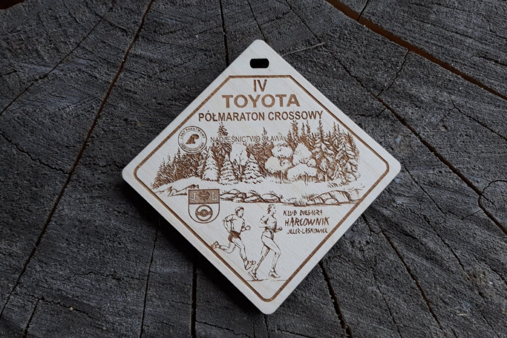 Drewniany medal w kształcie trapezu z grawerem górskiego szlaku, drzew, logotypów, napisem Półmaraton Crossowy i geometrycznymi detalami.