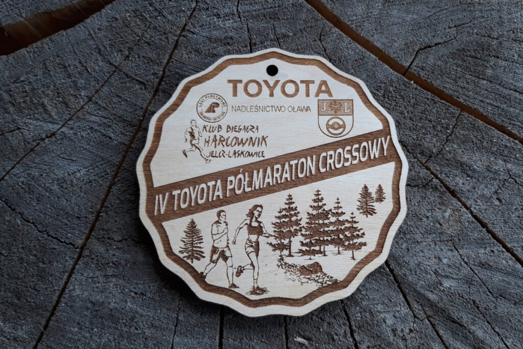 Drewniany medal o nieregularnych, pofalowanych brzegach z grawerem biegacza na leśnej ścieżce i napisem Półmaraton Crossowy.