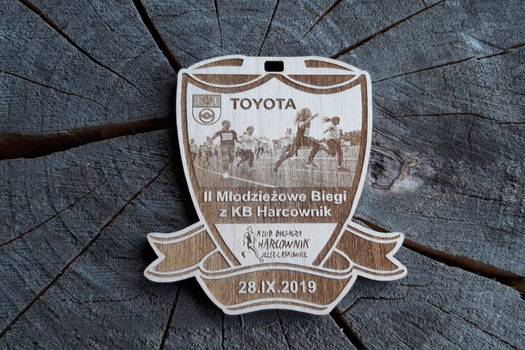 Drewniany medal w kształcie tarczy z wstęgą z wygrawerowanym zdjęciem biegnących dzieci, napisem II młodzieżowe Biegi z KB Harcownik, oraz logotypem Klubu.