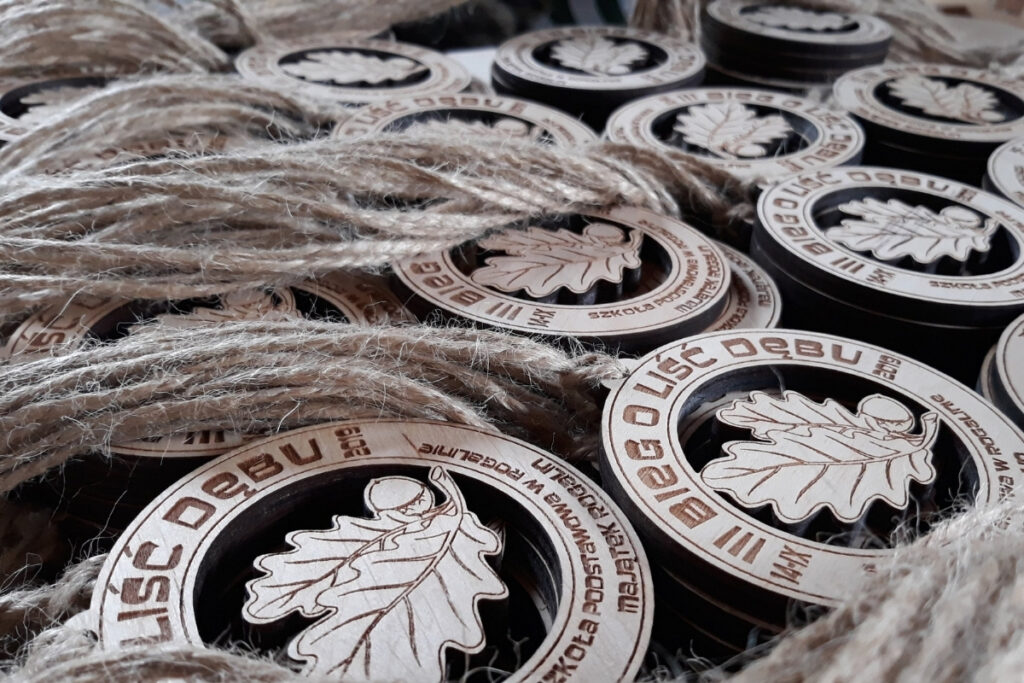 Okrągły drewniany medal z ażurowym, pustym wycięciem w kształcie liścia dębu i grawerem nazwy biegu.