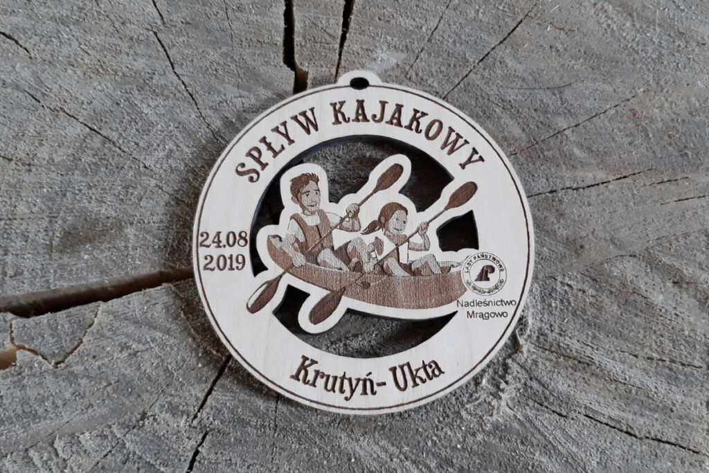 Okrągły drewniany medal z ażurowym wycięciem przedstawiającym dwóch kajakarzy w kajaku, grawerem fal oraz napisem Spływ Kajakowy.