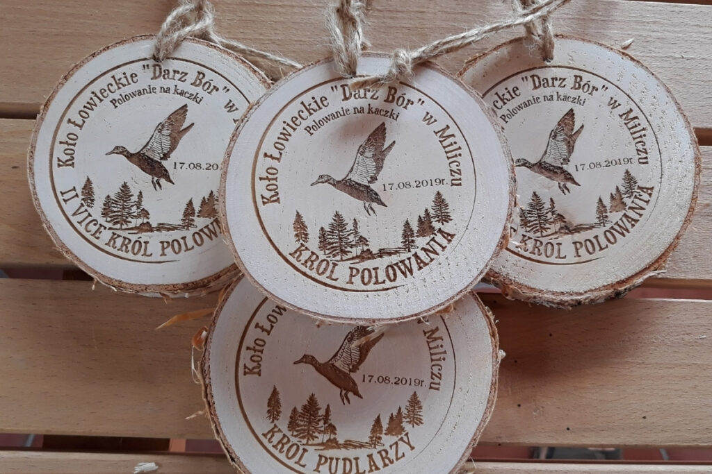 Medal z naturalnego plastra brzozy z korą, z grawerem kaczki krzyżówki zrywającej się do lotu, napisem Polowanie na Kaczki i ozdobnym motywem zalesienia.