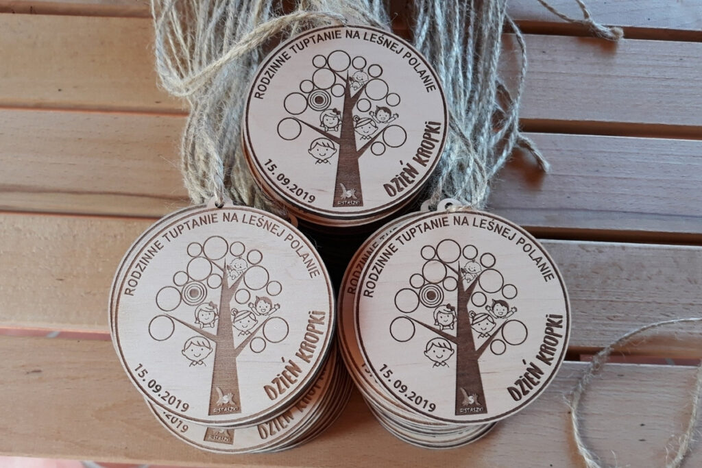 Okrągły drewniany medal z grawerem radosnej rodziny wśród gałęzi drzew, napisem Rodzinne Tuptanie na Leśnej Polanie i logotypem wydarzenia.