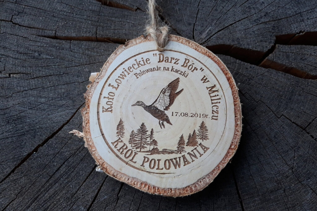 Medal z naturalnego plastra brzozy z korą, z grawerem kaczki krzyżówki zrywającej się do lotu, napisem Polowanie na Kaczki i ozdobnym motywem zalesienia.