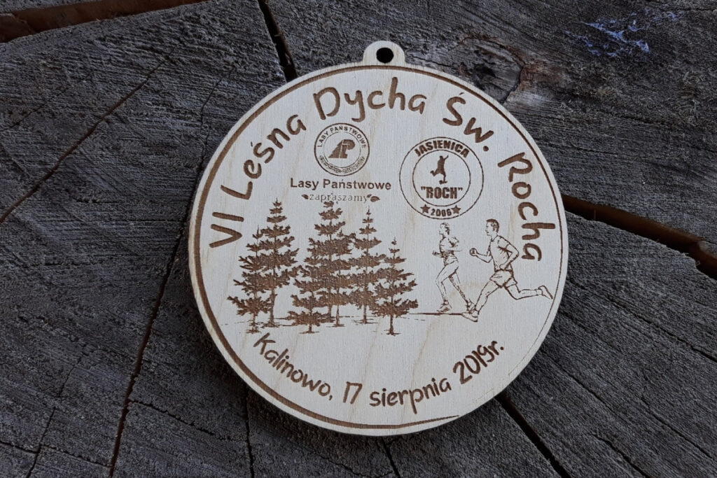 Okrągły drewniany medal z grawerem dwóch biegaczy na tle gęstego lasu iglastego, napisem VI Leśna Dycha św. Rocha i rokiem wydarzenia.