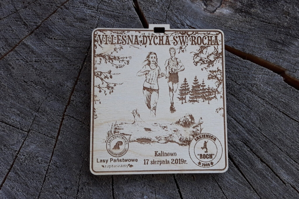Kwadratowy medal z drewna brzozowego z grawerem gęstego lasu, sylwetką biegacza i napisem Leśna Dycha.