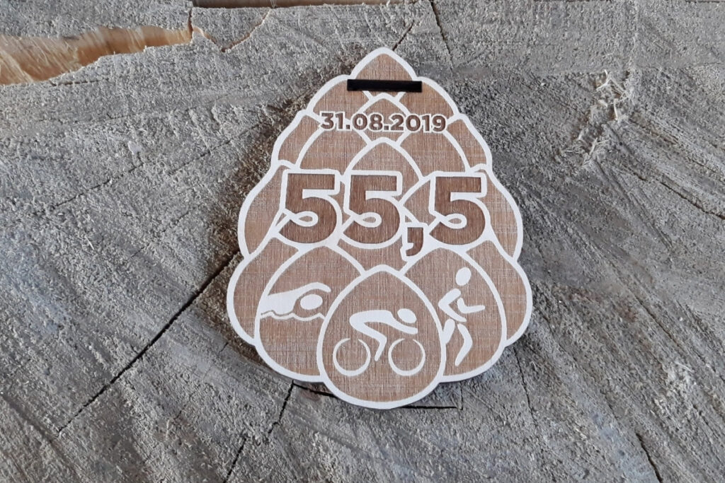 Drewniany medal w kształcie szyszki z wygrawerowanymi ikonami pływania, jazdy na rowerze i biegania oraz napisem Triathlon.