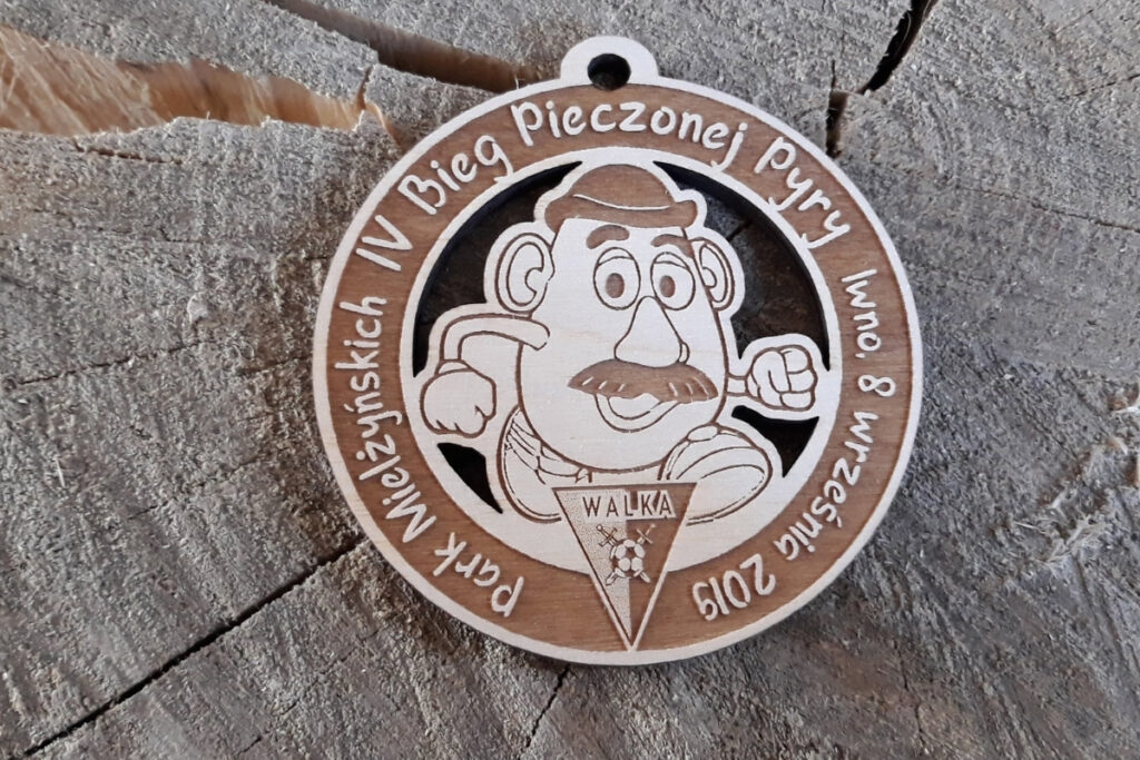 Okrągły drewniany medal z grawerem uśmiechniętego ziemniaka (pyry) w butach biegowych, napisem Bieg Pieczonej Pyry.