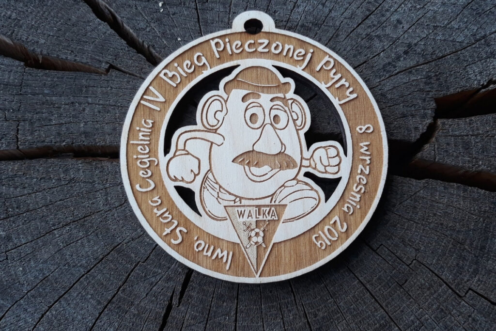 Okrągły drewniany medal z grawerem uśmiechniętego ziemniaka (pyry) w butach biegowych, napisem Bieg Pieczonej Pyry.