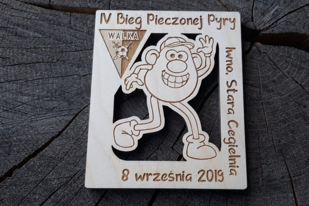 Kwadratowy drewniany medal z ażurowo wyciętą postacią biegnącego ziemniaka w środku oraz grawerem IV Bieg Pieczonej Pyry.