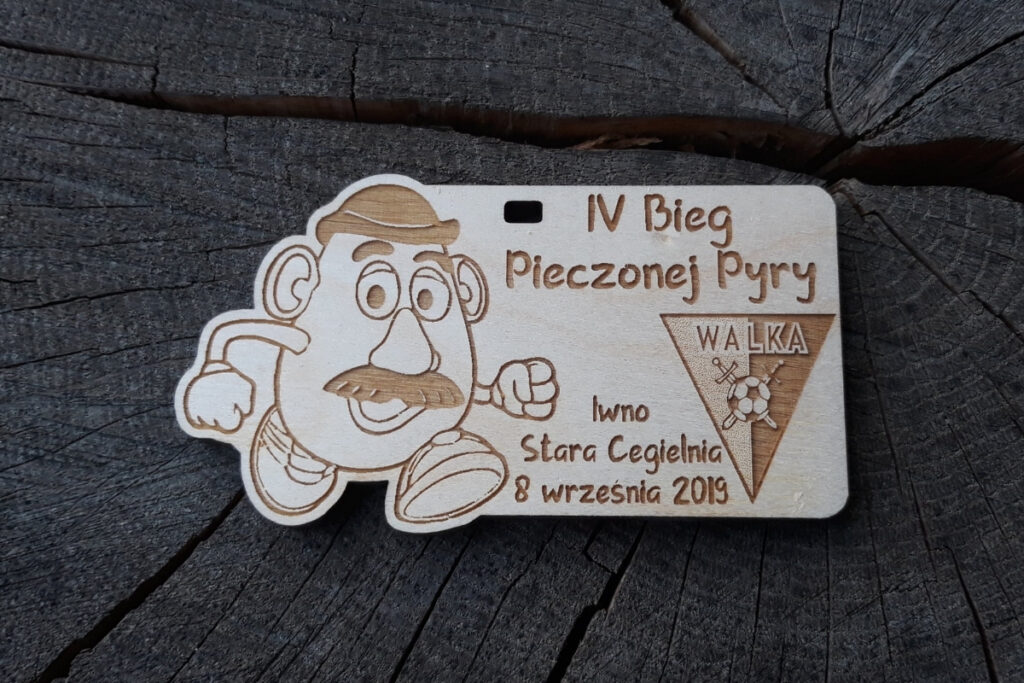 Kwadratowy drewniany medal z ażurowo wyciętą postacią biegnącego ziemniaka w środku oraz grawerem IV Bieg Pieczonej Pyry.